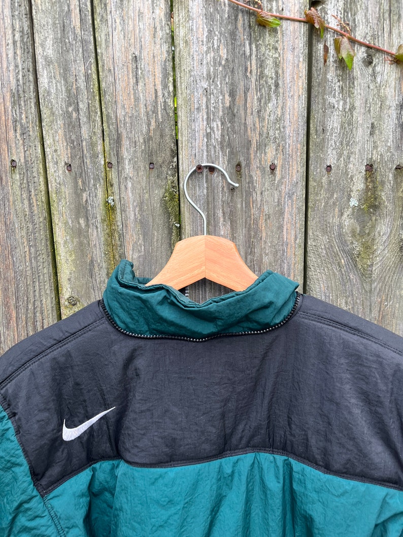 May include: A black and green jacket with a white Nike swoosh logo on the back. The jacket has a zipper closure and a high collar.