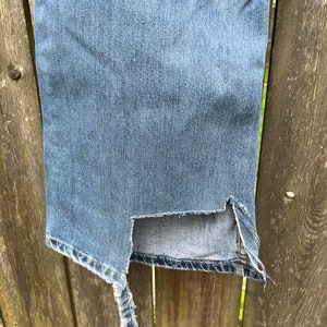 May include: A pair of blue denim jeans with a large rip on the side seam. The jeans are hanging on a wooden fence.