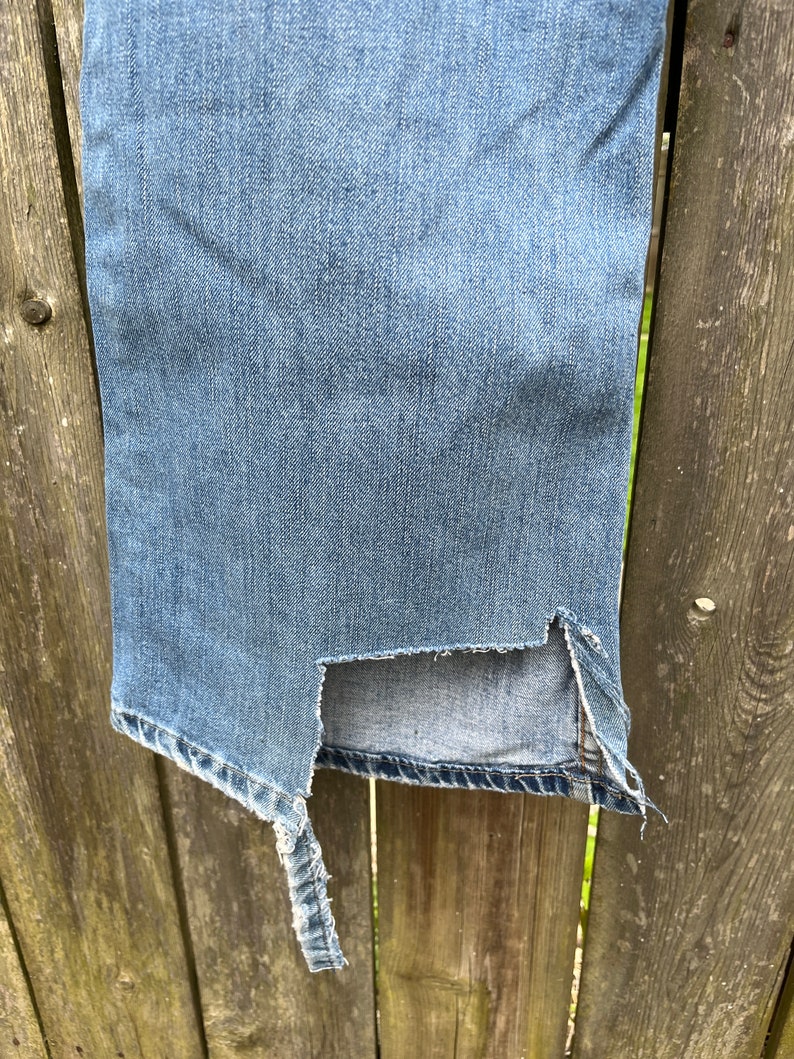 May include: A pair of blue denim jeans with a large rip on the side seam. The jeans are hanging on a wooden fence.