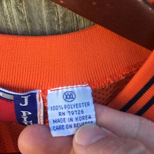 K&ouml;nnte beinhalten: Nahaufnahme eines wei&szlig;en Etiketts mit schwarzem Text, der "100% POLYESTER RN 79726 MADE IN KOREA CARE ON REVERSE" lautet, mit einem kleinen wei&szlig;en Etikett mit schwarzem Text, das "XXL" lautet, das oben angebracht ist. Das Etikett ist an einem orangefarbenen Netzshirt befestigt.