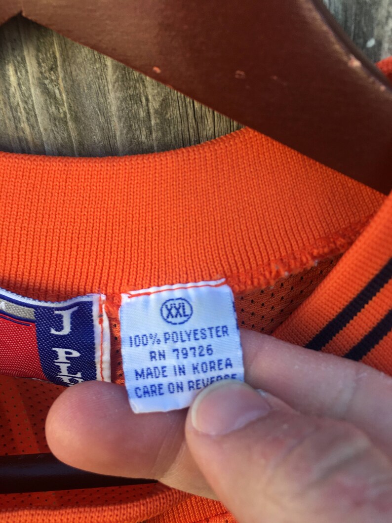 K&ouml;nnte beinhalten: Nahaufnahme eines wei&szlig;en Etiketts mit schwarzem Text, der "100% POLYESTER RN 79726 MADE IN KOREA CARE ON REVERSE" lautet, mit einem kleinen wei&szlig;en Etikett mit schwarzem Text, das "XXL" lautet, das oben angebracht ist. Das Etikett ist an einem orangefarbenen Netzshirt befestigt.