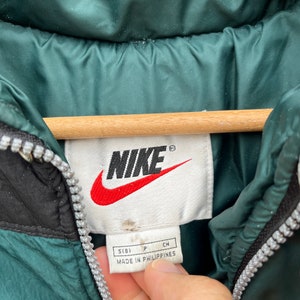 May include: A green Nike puffer jacket with a black zipper and a white tag that says "Nike" in black lettering and "Made in Philippines" in black lettering. The tag also has the size "S(8)" in black lettering.