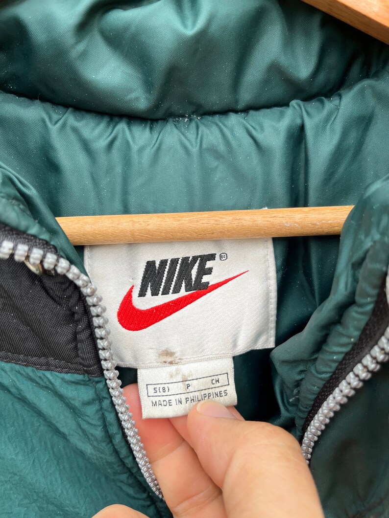 May include: A green Nike puffer jacket with a black zipper and a white tag that says "Nike" in black lettering and "Made in Philippines" in black lettering. The tag also has the size "S(8)" in black lettering.