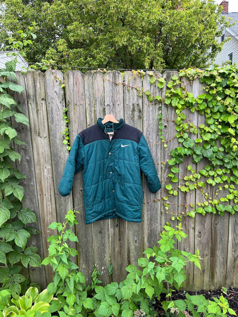 May include: A dark green and black quilted jacket with a white Nike logo on the left chest. The jacket has a button closure and a drawstring at the waist.