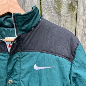 May include: A green and black Nike jacket with a white swoosh logo. The jacket has a zipper closure and a button on the front.