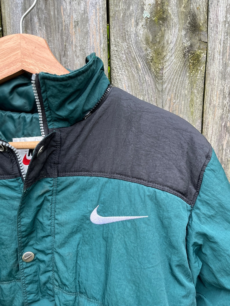 May include: A green and black Nike jacket with a white swoosh logo. The jacket has a zipper closure and a button on the front.