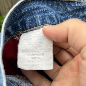 May include: A white tag with black text attached to a pair of blue jeans. The tag reads "MADE IN BANGLADESH", "100% COTTON", "98RYWEM", "36X30", and "SAM".