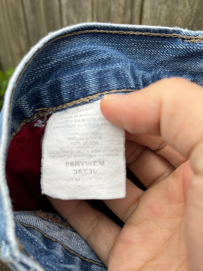 May include: A white tag with black text attached to a pair of blue jeans. The tag reads "MADE IN BANGLADESH", "100% COTTON", "98RYWEM", "36X30", and "SAM".