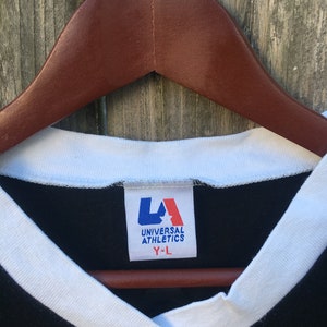 May include: Close-up of a black V-neck t-shirt with white trim and a tag that reads "UNIVERSAL ATHLETICS Y-L". The shirt is hanging on a brown wooden hanger against a weathered wood background.