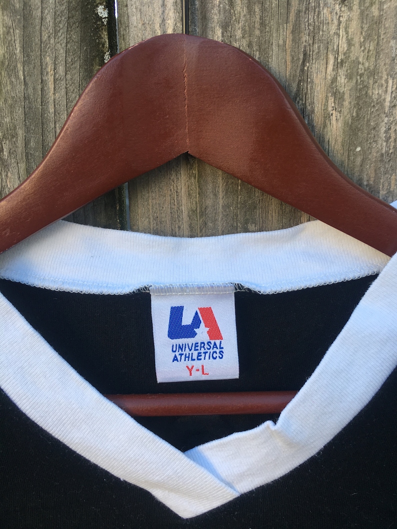 May include: Close-up of a black V-neck t-shirt with white trim and a tag that reads "UNIVERSAL ATHLETICS Y-L". The shirt is hanging on a brown wooden hanger against a weathered wood background.