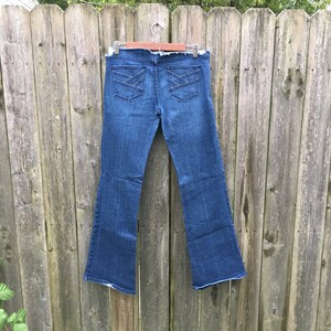 May include: Dark wash denim jeans with a flared leg design. The jeans feature two back pockets with decorative stitching. The jeans are hanging on a wooden hanger against a weathered wooden fence.