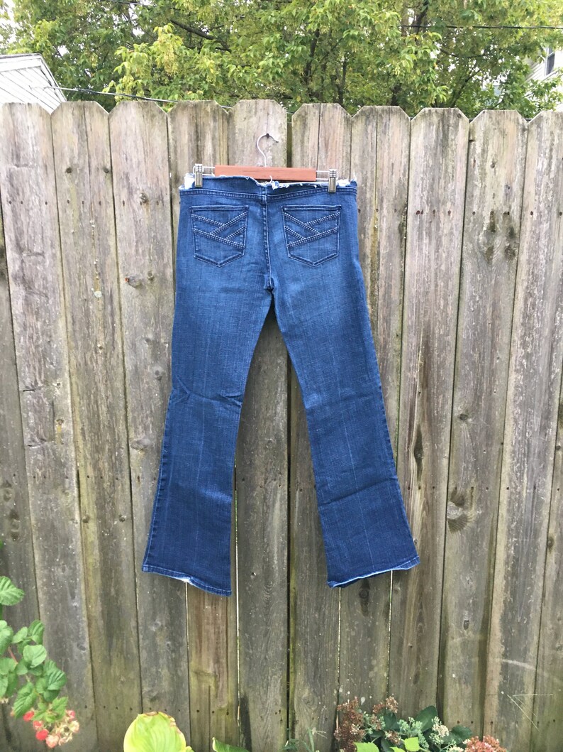 May include: Dark wash denim jeans with a flared leg design. The jeans feature two back pockets with decorative stitching. The jeans are hanging on a wooden hanger against a weathered wooden fence.