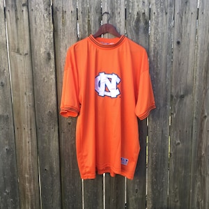 K&ouml;nnte beinhalten: Orangefarbenes T-Shirt mit einem wei&szlig;en "NC"-Logo und schwarzem Besatz. Das Shirt h&auml;ngt an einem Holzb&uuml;gel vor einem Holzzaun.