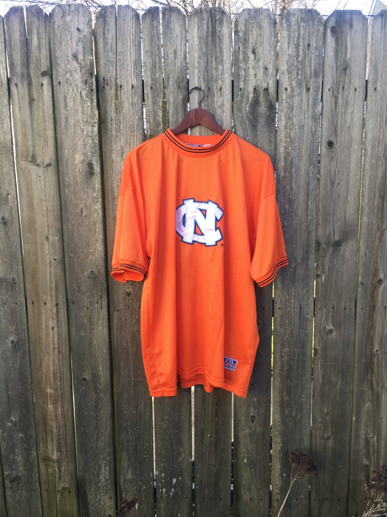 K&ouml;nnte beinhalten: Orangefarbenes T-Shirt mit einem wei&szlig;en "NC"-Logo und schwarzem Besatz. Das Shirt h&auml;ngt an einem Holzb&uuml;gel vor einem Holzzaun.