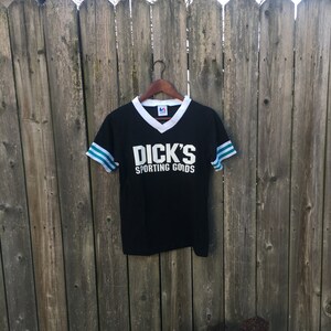 May include: Black v-neck t-shirt with white collar and sleeve trim, featuring the text "DICK'S SPORTING GOODS" in white block letters. The sleeves have teal and white stripes. The shirt hangs on a wooden hanger.