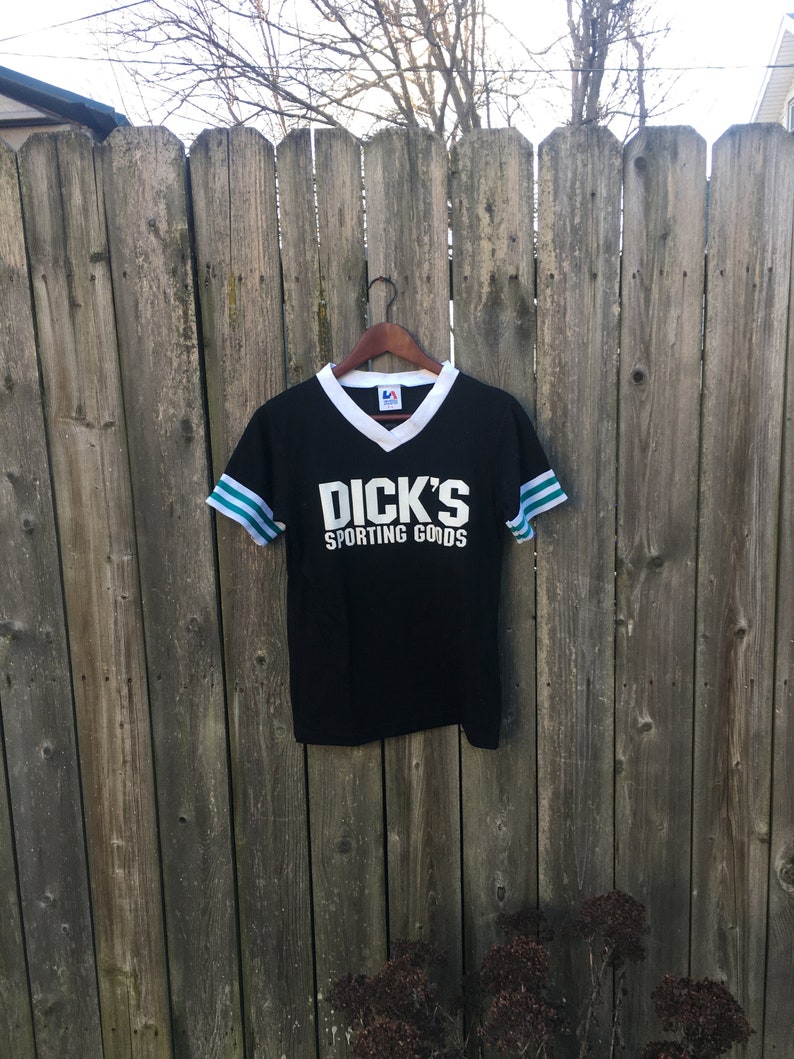 May include: Black v-neck t-shirt with white collar and sleeve trim, featuring the text "DICK'S SPORTING GOODS" in white block letters. The sleeves have teal and white stripes. The shirt hangs on a wooden hanger.