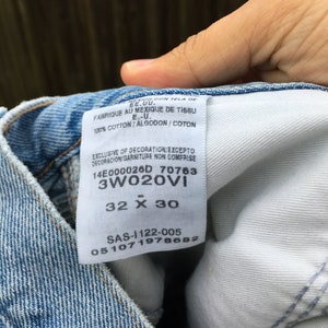 May include: A white tag sewn into a pair of blue denim jeans. The tag has the text "14E000026D 70763 3W020VI 32 X 30 SAS-1122-005 051071978682" printed on it.