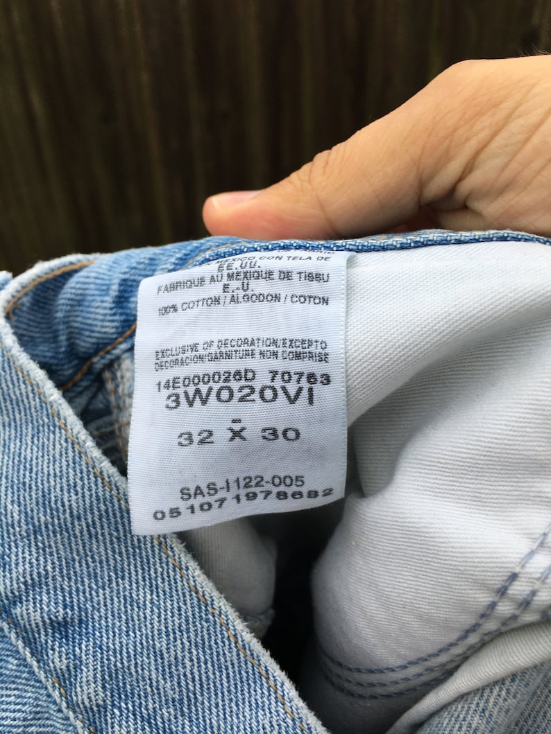 May include: A white tag sewn into a pair of blue denim jeans. The tag has the text "14E000026D 70763 3W020VI 32 X 30 SAS-1122-005 051071978682" printed on it.