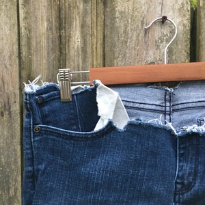 May include: Dark blue denim jeans with a raw, frayed hem and pocket detail. The jeans are hanging on a wooden hanger against a weathered wooden fence.