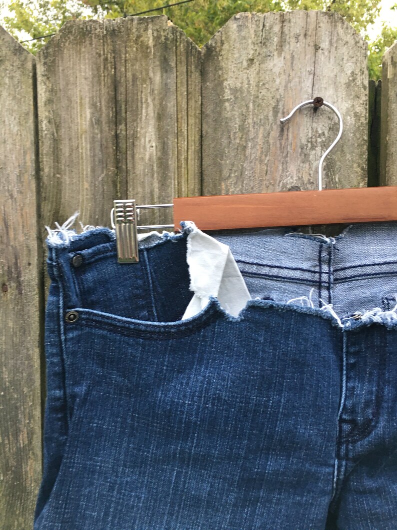 May include: Dark blue denim jeans with a raw, frayed hem and pocket detail. The jeans are hanging on a wooden hanger against a weathered wooden fence.