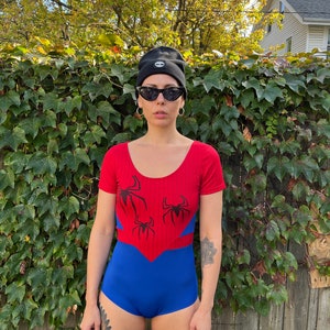 Pu&ograve; includere: Un costume da bagno rosso e blu con un design a ragnatela e tre ragni neri. Il costume da bagno ha maniche corte e una scollatura rotonda.