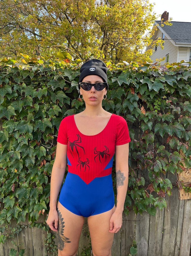 Pu&ograve; includere: Un costume da bagno rosso e blu con un design a ragnatela e tre ragni neri. Il costume da bagno ha maniche corte e una scollatura rotonda.