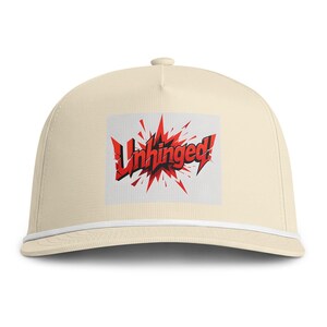 May include: A cream-colored baseball cap featuring a white cord trim along the base of the crown. The front panel displays a graphic with the word Unhinged! in bold black letters set against a jagged, explosive red and black comic book style burst.