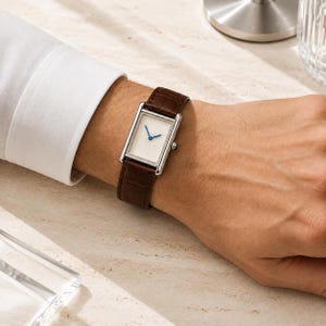 Op de afbeelding: Een rechthoekig polshorloge met een zilverkleurige kast en een witte wijzerplaat met een zonnestraalpatroon. Het horloge heeft twee dunne blauwe wijzers en is bevestigd met een donkerbruine leren band met een textuur en reliëfafwerking.