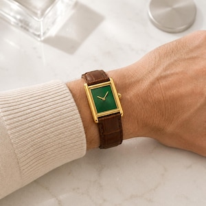 Op de afbeelding: Een rechthoekig polshorloge met een goudkleurige kast, een diepgroene wijzerplaat en een bruine lederen band met een gestructureerd krokodilpatroon. Het horloge heeft dunne goudkleurige wijzers en een kleine goudkleurige kroon aan de zijkant.