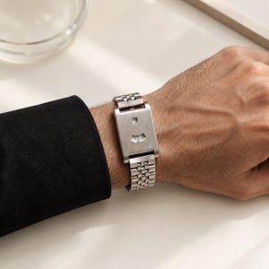 May include: A silver-toned rectangular wristwatch featuring a brushed metal finish, a link bracelet, and a unique dial design with two small horizontal windows displaying numerical time indicators, worn with a black long-sleeved garment.