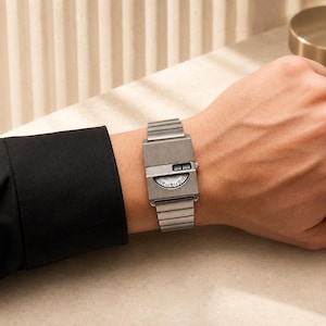 Peut inclure: Une montre-bracelet rectangulaire argentée avec une finition en métal brossé et un bracelet à maillons assorti. Le cadran présente une forme semi-circulaire avec des chiffres et une petite fenêtre numérique affichant l'heure en noir.