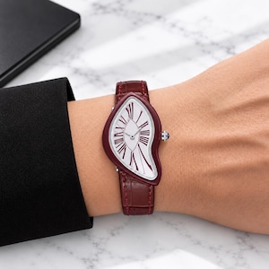 Peut inclure: Une montre-bracelet dotée d'un boîtier asymétrique en forme de goutte d'un fini bordeaux profond. Le cadran blanc présente des chiffres romains noirs allongés et des aiguilles argentées. Elle est assortie d'un bracelet en cuir gaufré croco.