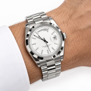 Op de afbeelding: Een zilverkleurig roestvrijstalen polshorloge met een witte wijzerplaat, zilveren streepindexen en zilveren wijzers. Het horloge heeft een dagweergave op de twaalf uur positie, een datumvenster op drie uur en een schakelarmband.