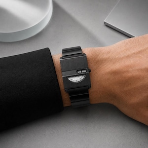 Könnte beinhalten: Eine mattschwarze Armbanduhr mit rechteckigem Gehäuse und passendem Gliederarmband. Das Zifferblatt verfügt über eine halbkreisförmige Anzeige mit weißen Markierungen und ein kleines digitales Fenster, das die Zahlen 12 und 00 horizontal anzeigt.