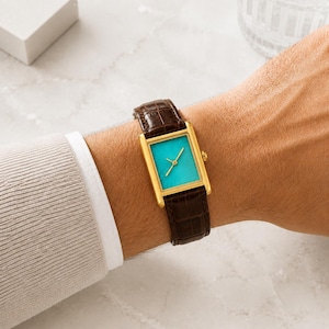Op de afbeelding: Een rechthoekig polshorloge met een goudkleurige kast en een levendige turquoise wijzerplaat met zonnestraalpatroon. Het horloge heeft dunne gouden wijzers en een donkerbruine leren band met een textuur van krokodillenleer.