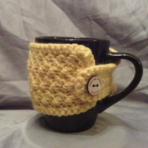 May include: A black coffee mug with a crocheted cozy in a light yellow color. The cozy has a button closure and a label that reads "Dancing Stitches Crochet". The cozy is wrapped around the mug.