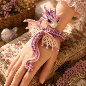 May include: A crochet dragon bracelet featuring a purple, pink, and cream color palette. The design includes a detailed dragon head, intricate lace-like wings, and a long tail that extends down to a star-shaped ring, accented with small white pearls.