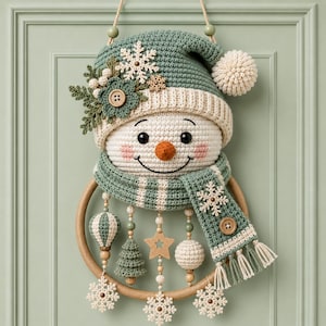 May include: A crocheted snowman wall hanging featuring a sage green hat with a white pom-pom, a matching scarf, and a wooden ring base. The snowman is decorated with crocheted snowflakes, a wooden star, a small tree, and dangling wooden beads.