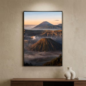 Pode incluir: Uma impressão de arte de parede vertical emoldurada retratando uma paisagem vulcânica ao nascer do sol. A cena apresenta uma montanha cônica proeminente em primeiro plano com encostas texturizadas, cercada por névoa baixa e um pico distante.