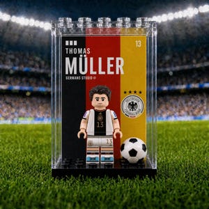 Puede incluir: Una minifigura de Thomas Müller con una camiseta de fútbol alemana blanca y negra con el número 13, exhibida en una caja de plástico transparente. El fondo muestra los colores de la bandera alemana, el texto "THOMAS MÜLLER" y un balón.