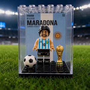 Könnte beinhalten: Eine Diego Maradona Minifigur im Argentinien-Trikot mit der Nummer 10, präsentiert in einer durchsichtigen Kunststoffbox mit einem Fußball und einem goldenen Pokal. Der Hintergrund zeigt den Namen Diego Maradona und ein Sonnenemblem.