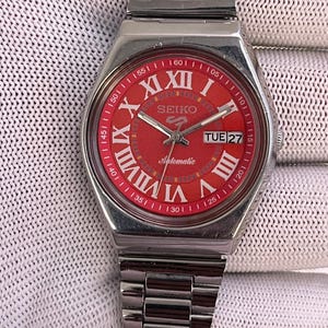Peut inclure: Une montre-bracelet automatique Seiko 5 en acier inoxydable argenté avec un cadran rouge vif. Le cadran présente de grands chiffres romains blancs, un guichet jour-date à trois heures et des aiguilles argentées. Bracelet à maillons assorti.