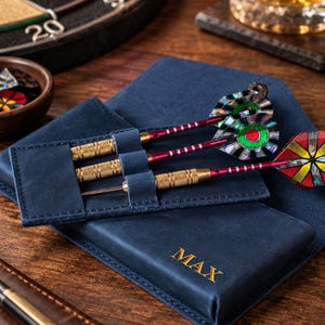May include: A navy blue leather dart case featuring the name MAX embossed in gold lettering. The case holds three darts with gold-toned textured barrels, red shafts, and iridescent multi-colored flights. The case has individual slots for each dart.