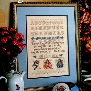 May include: A cross-stitch pattern featuring the alphabet, a quote about angels, and three angels with wings. The quote reads "Be not forgetful to entertain strangers: for thereby some have entertained angels unawares. Heb. 13:2". The pattern is framed and displayed on a table with a teapot and a small box with an angel design.