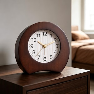Puede incluir: Un reloj de mesa redondo de madera marrón oscuro con una esfera blanca de rayas verticales. El reloj tiene números arábigos negros del 1 al 12 y manecillas doradas para las horas, minutos y segundos. El marco tiene un diseño curvo.
