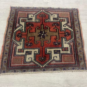 May include: A square hand-woven rug featuring a central geometric motif in shades of rust red, tan, and cream. The design includes intricate angular patterns and floral-like accents, framed by a decorative border with fringe on the edges.