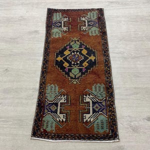 May include: A rectangular woven rug featuring a deep rust-orange background with a central dark navy blue diamond medallion. The design includes geometric motifs in light green, yellow, and white, framed by a dark border with intricate patterns.