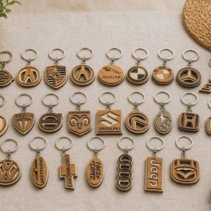 May include: A collection of twenty-three wooden keyrings featuring various automotive brand logos and emblems. Each keyring is crafted from light-toned wood, includes a silver-coloured metal split ring, and displays distinct laser-cut designs.