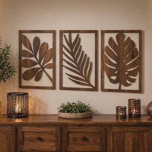 May include: Three rectangular wooden wall art panels featuring dark brown laser-cut leaf silhouettes. The designs include a multi-lobed leaf, a long palm frond, and a large monstera leaf, all mounted within simple frames against a light wall.