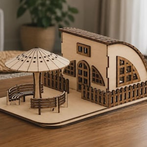 May include: A laser-cut wooden model of a small house with a curved roof, featuring a fenced yard, a central door, and arched windows. A matching wooden patio set with a circular umbrella and two curved benches sits on the base platform.